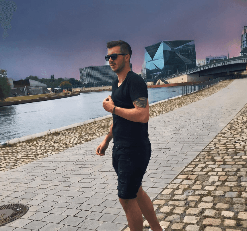 Man wearing sunglasses and black clothing walking along a riverside path with modern buildings in the background.